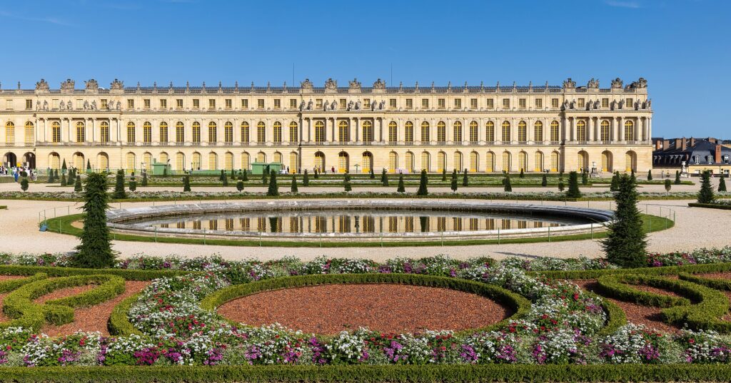Garden façade of the Palace of Versailles with the Parterre du Midi flower beds and pond under a blue sky